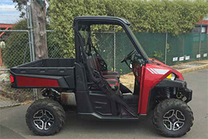 This image shows a small red truck, side on. There is a tray back. The driver’s cabin has no doors, seats for two people, and a steering wheel. It is a four-wheeled vehicle but in this view only two are seen.